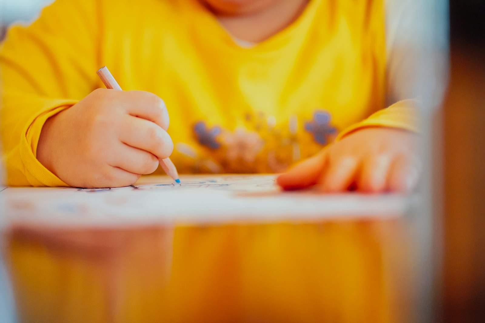 A child's hand carefully coloring with a pencil at a table, wearing a warm yellow sweater.